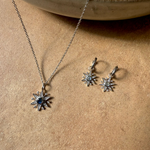 Silver sun-shaped necklace and earrings on a beige surface with a ceramic bowl in the background.