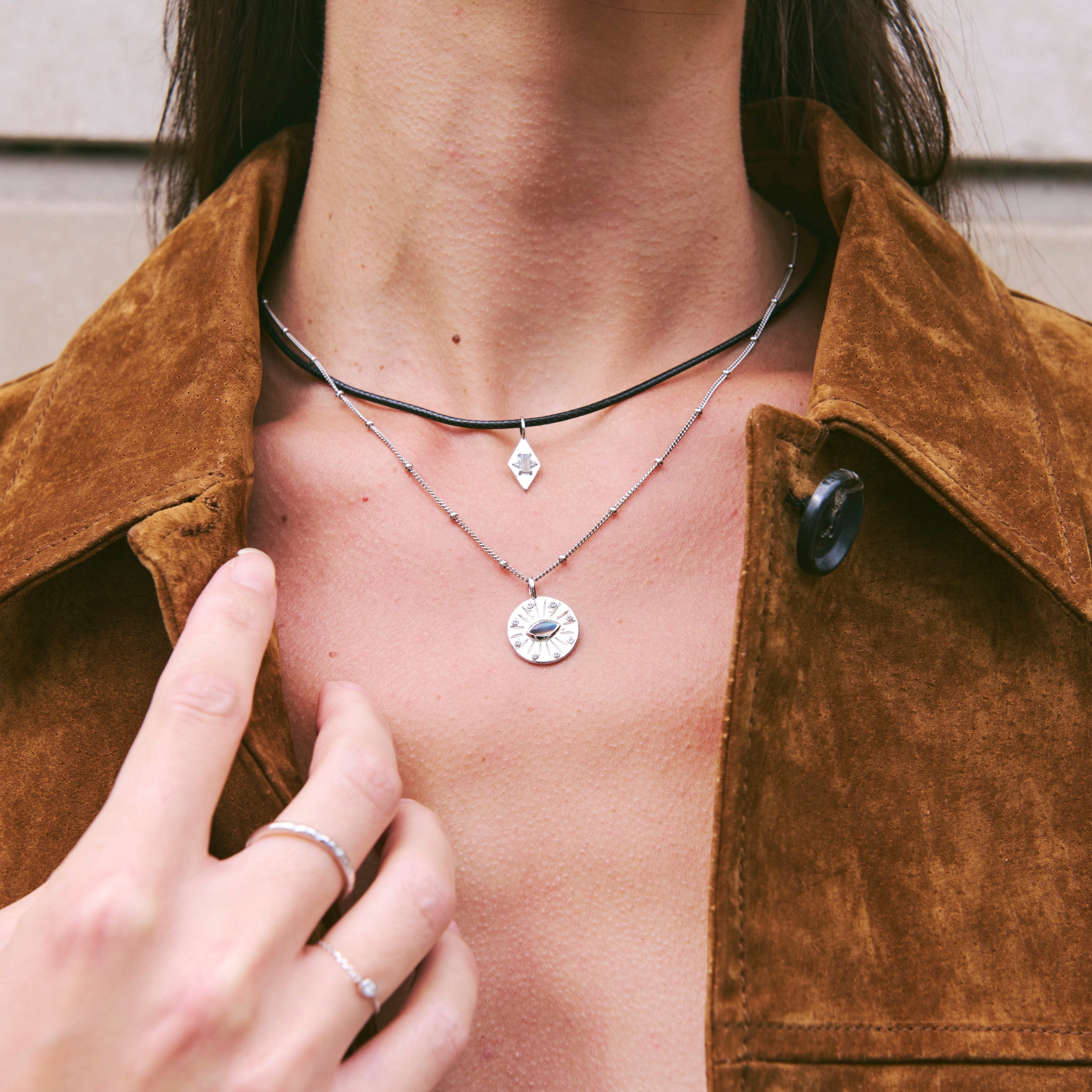 Person wearing a necklace with a pendant, pointing to it, against a neutral background.